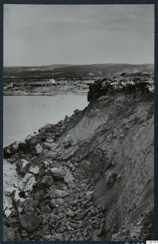 Eroded cliff edge with loose rocks at its foot, calm water on the left, and low hills and a small white building in the distance.