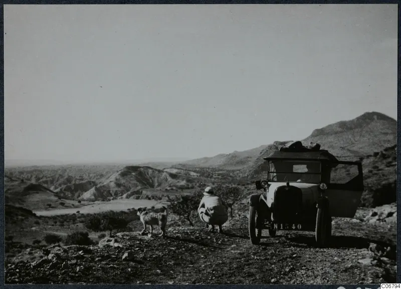 Landscape from the road to Hallefka, with a parked car, a crouching person and a dog on stony ground.