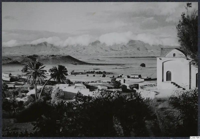 Village of flat-roofed houses and palm trees on a plain, with mountains behind and a whitewashed building at right