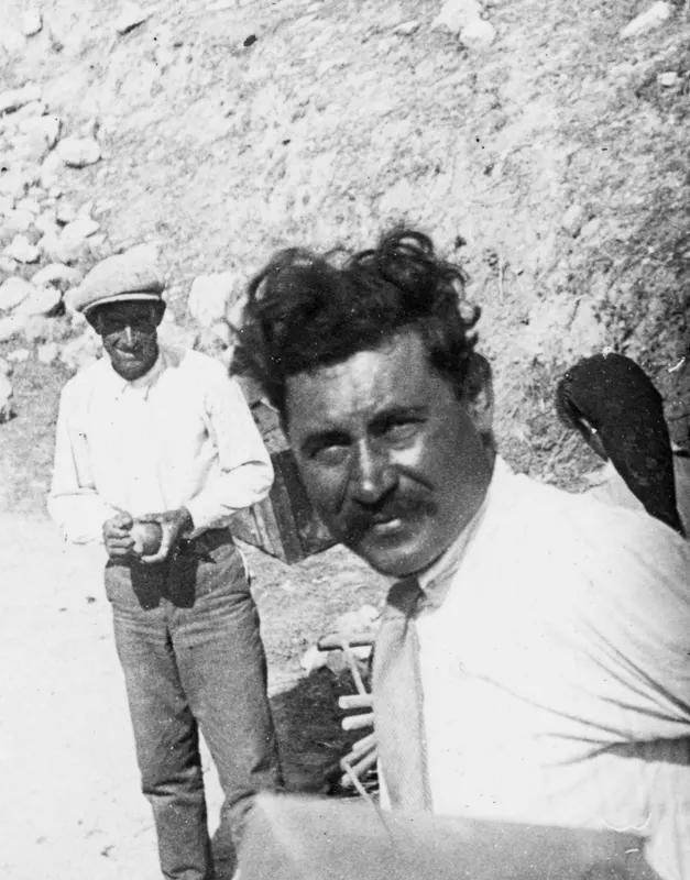 Two men at Mersinaki in front of an earth bank with stones; one wears a flat cap and holds a small object.