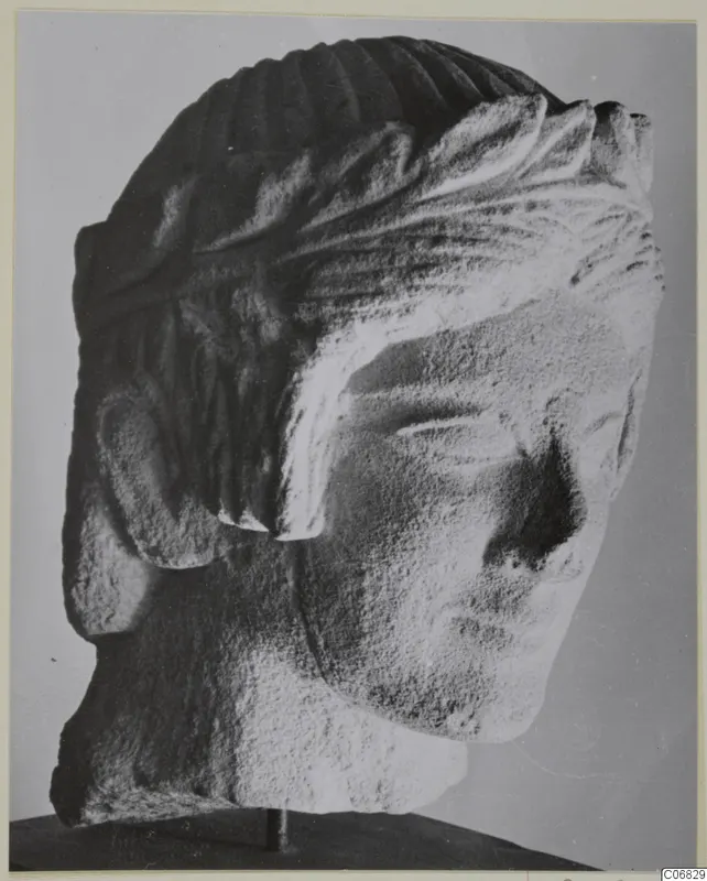 Limestone head from Mersinaki with carved hair in grooves and locks, shown on a dark stand