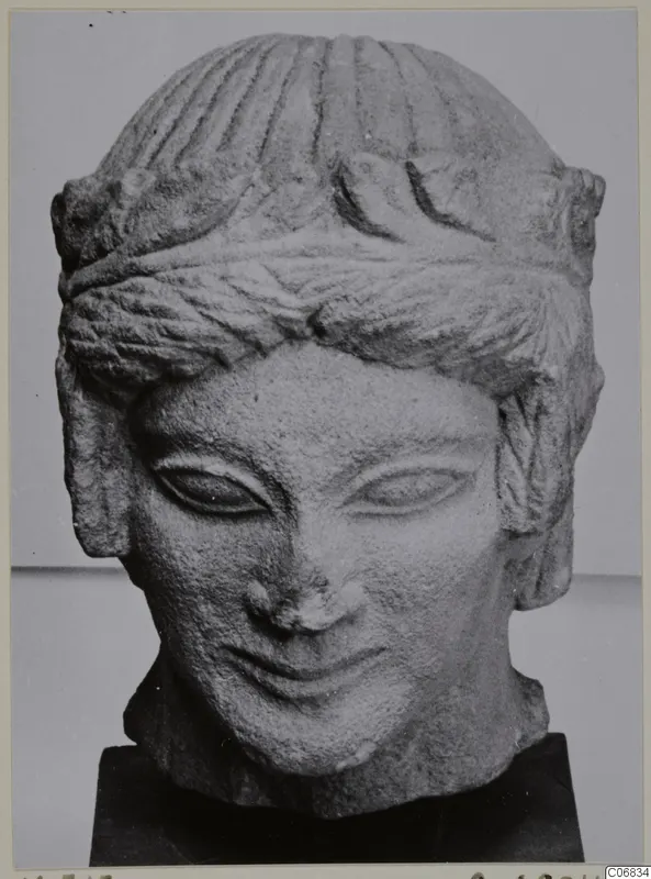 Carved limestone head with a bust-like neck and banded hair, shown on a black stand from Mersinaki