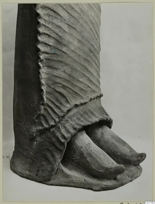 Lower part of a terracotta figure with a pleated skirt and two pointed feet, photographed close up against a plain background.