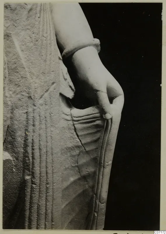 Close view of a limestone statue fragment showing a bare arm with a bracelet, the hand at the hip, and carved drapery folds.