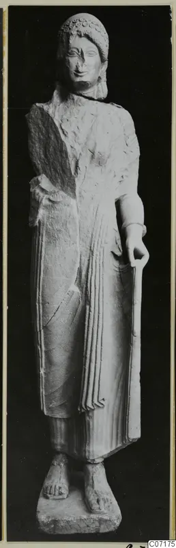 Limestone statue of a standing woman from Vouni, holding a long staff, with a broken left side and worn face.