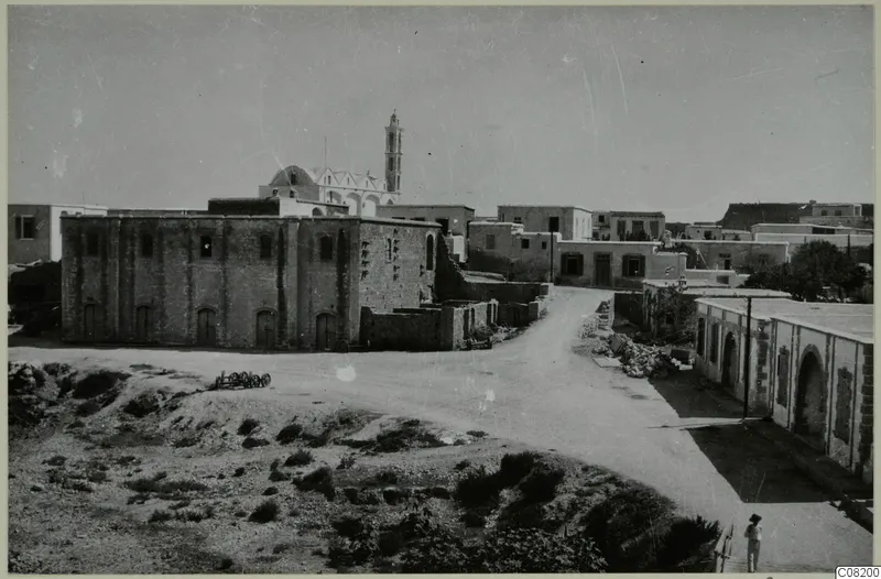 Street in Kyrenia with a large stone building, low houses, shrubs and one person standing at lower right.