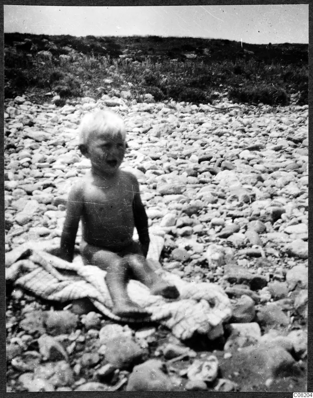 A small fair-haired child sitting on a blanket among rounded stones on open ground.
