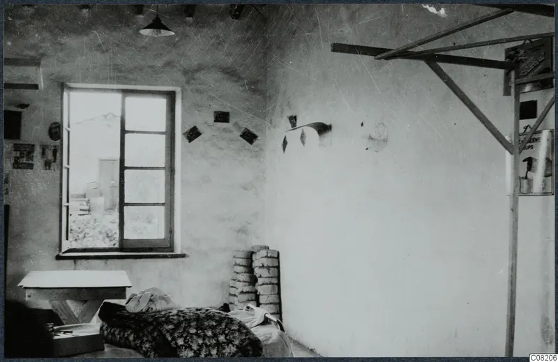 Interior of a plastered room with a window, a wooden table and a bed covered by a floral blanket.