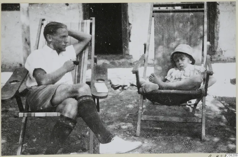 A man and a small child seated in wooden deck chairs outside a whitewashed building at Machairas.