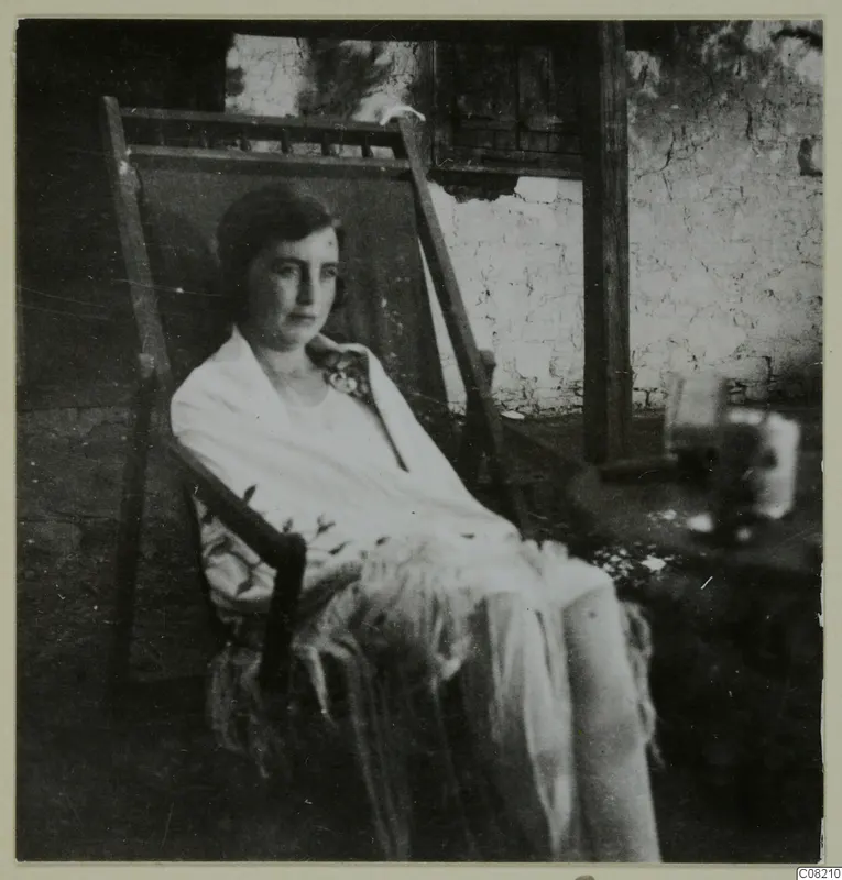 Woman seated in a wooden deck chair, wearing a light shawl, in front of a plastered wall.