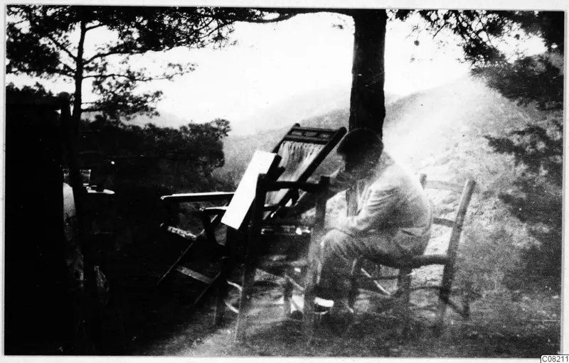 A man seated on a wooden chair under a pine tree, bent over a sheet of paper on a folding stand.