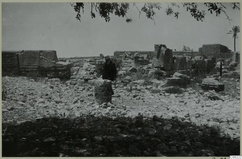 Man in a hat and dark coat standing among stone ruins, with a cylindrical column drum in the foreground