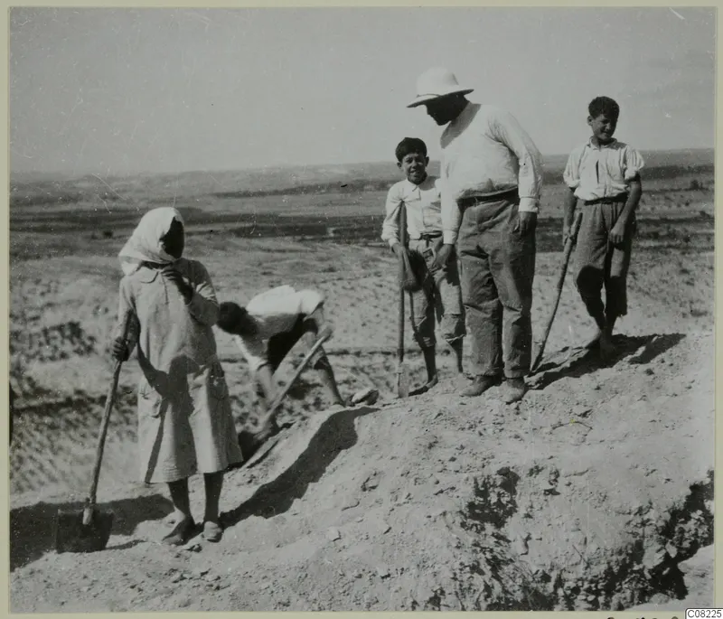 Five people with digging tools at the edge of an excavation, including a man in a pith helmet, with open hills behind.