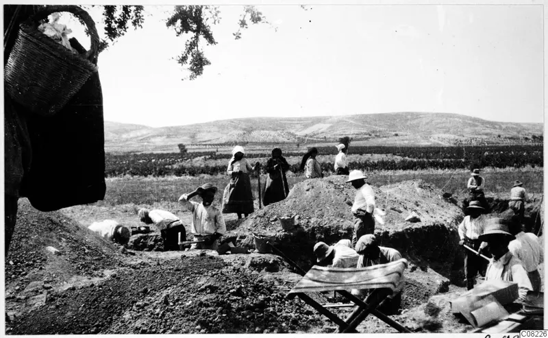Workers digging in an excavation trench, with a large basket hanging from a tree in the foreground and a folding stool at lower right