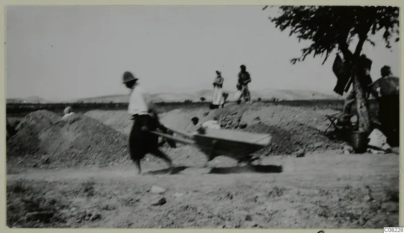 A blurred man pushes a wheelbarrow past earth heaps, with women standing nearby and low hills behind