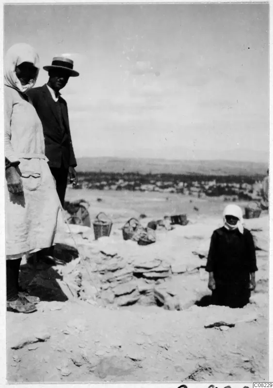 Woman, man in a straw boater and a smaller figure beside a stone wall in an excavated area, with baskets and a town below distant hills
