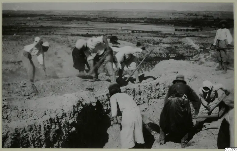 Workers digging a trench at an excavation, with a flat plain and low hills in the background.