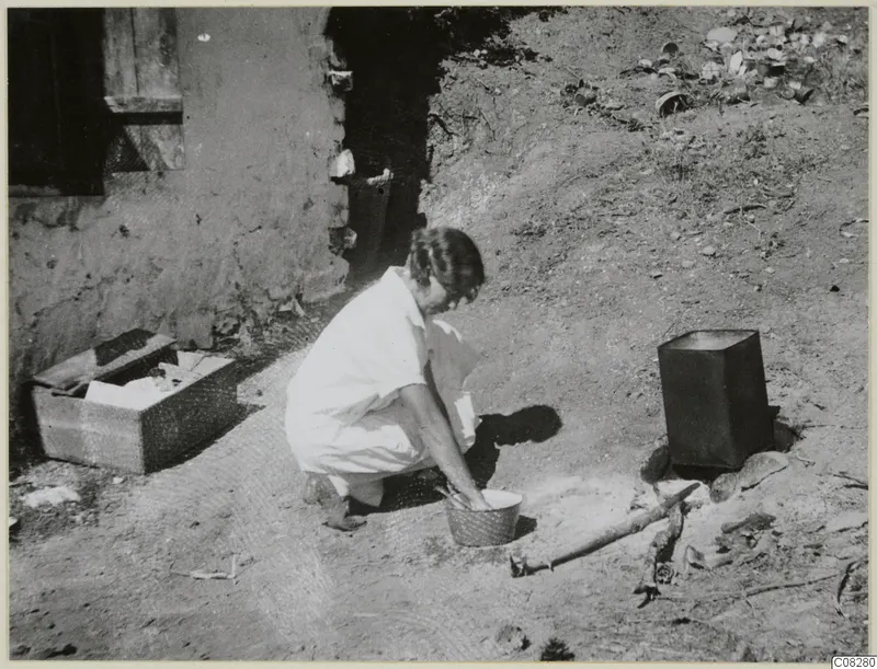 Woman crouching beside a round bowl on bare ground, apparently washing, outside a mud-walled building at Machairas