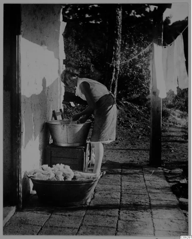 Woman washing cloth in a metal tub outside a whitewashed building, with a large tub of white laundry in front
