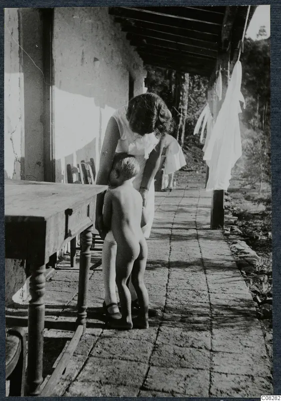 A woman bending over a small naked child on a brick-paved veranda beside a wooden table