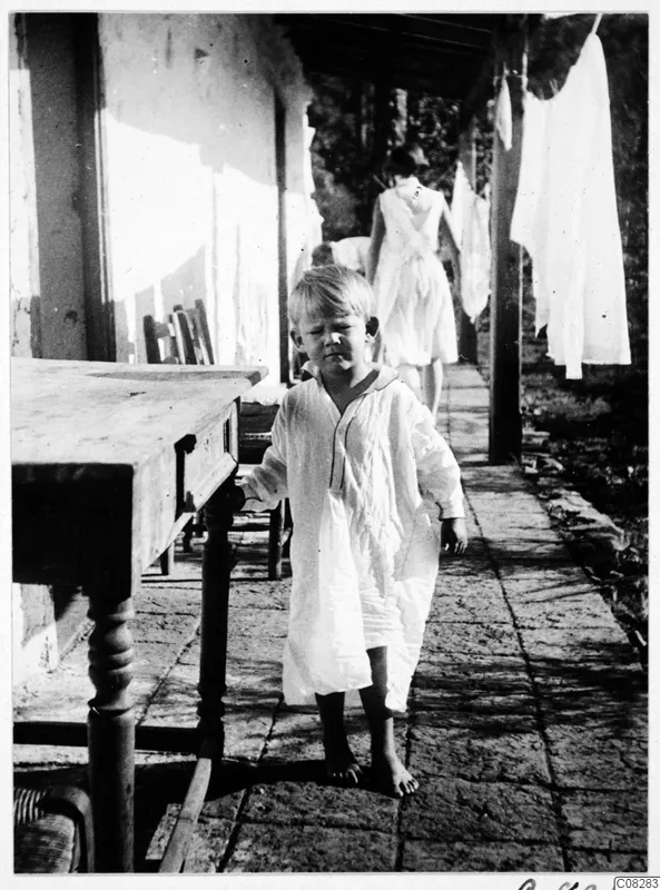 Small barefoot boy in a long white nightshirt beside a wooden table on a paved veranda with laundry hanging nearby.