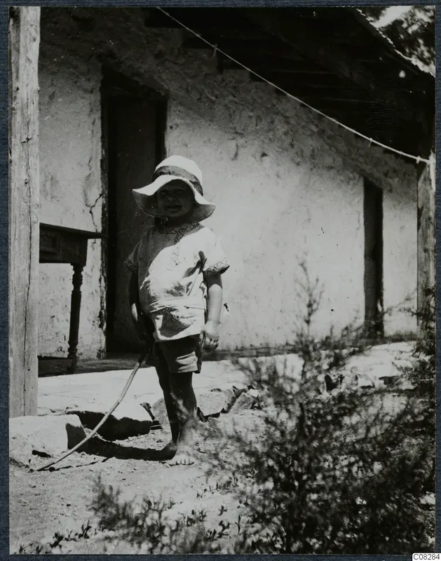 Young child in a wide-brimmed hat standing barefoot in front of a plastered building at Machairas