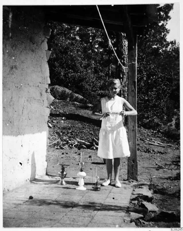 Woman in a light dress standing beside three oil lamps on a paved terrace next to a plastered mud wall.