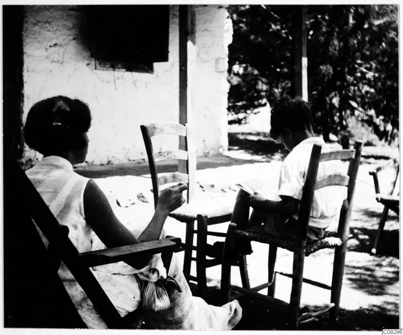 Two people sitting on a shaded veranda beside a whitewashed building, one reading, with wooden chairs around them.