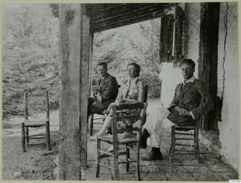 Three people seated on the veranda of a whitewashed building at Machairas, with rush-seated chairs around them.