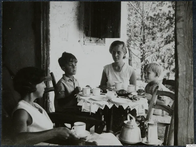 Four people seated at a table with cups, plates and a teapot on a shaded veranda beside a white wall