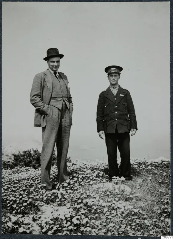 Two men standing in a field of small white flowers at Vouni, one in a suit and hat, one in a uniform cap.