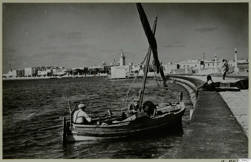 Wooden boat with three men in calm water beside a curved stone quay, with a town of white buildings along the shore
