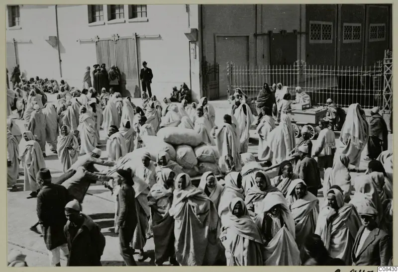 Crowd of people in pale hooded cloaks carrying sacks across a paved yard beside a large building