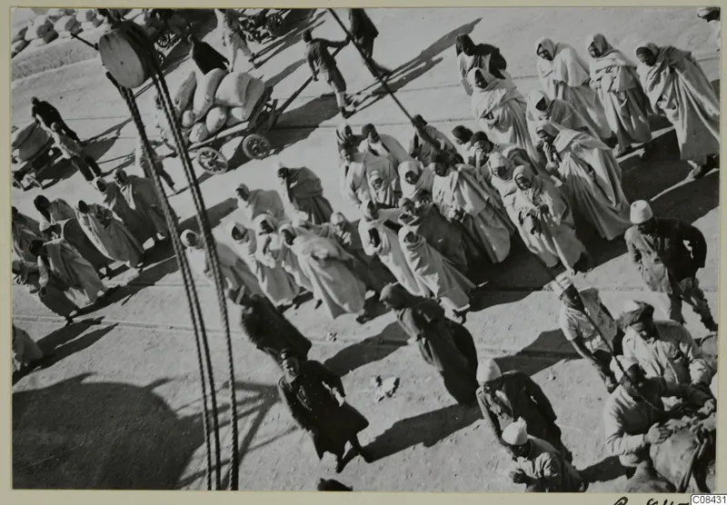 Crowd of people in long pale hooded robes walking across open ground, with hand carts and sacks and ropes in the foreground