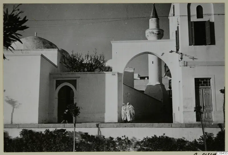 Whitewashed buildings with a dome, a tall tower and an arched passage, with two people in white robes walking past a hedge.