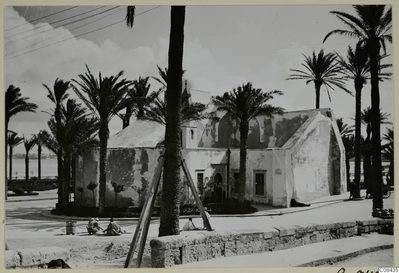 Whitewashed building with arched openings among palm trees, with people seated on a street corner, North Africa.