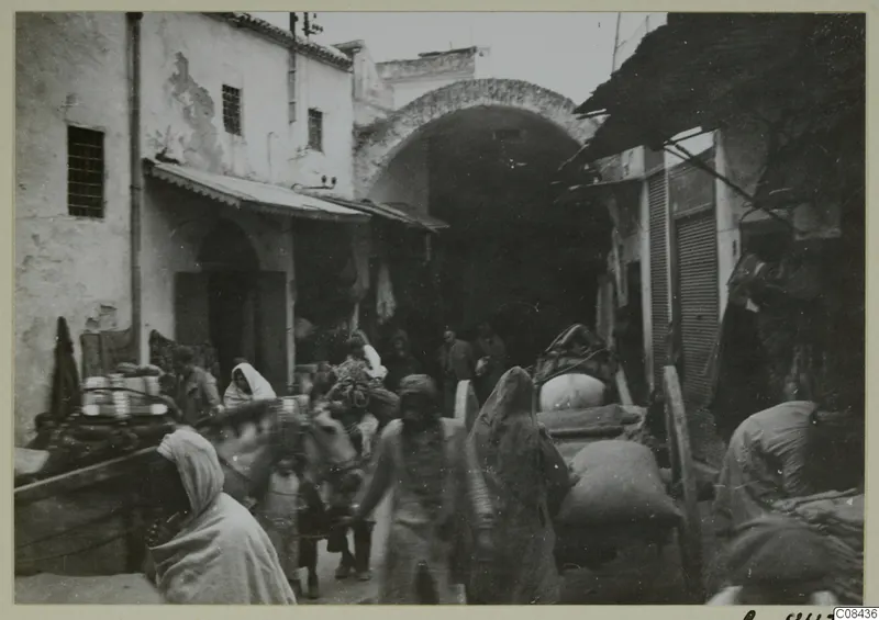 Street scene in North Africa with an arched passage, people, and a loaded donkey or mule.