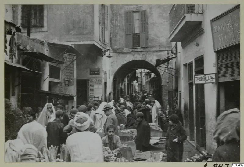 Crowded market street under an archway, with people and produce on low stalls, recorded as North Africa.