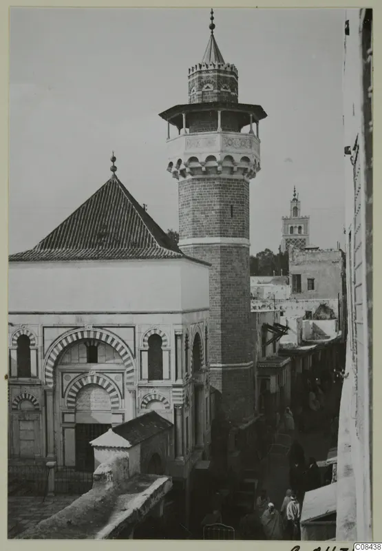 Mosque with a tall minaret and banded arches, with people in a narrow street, North Africa