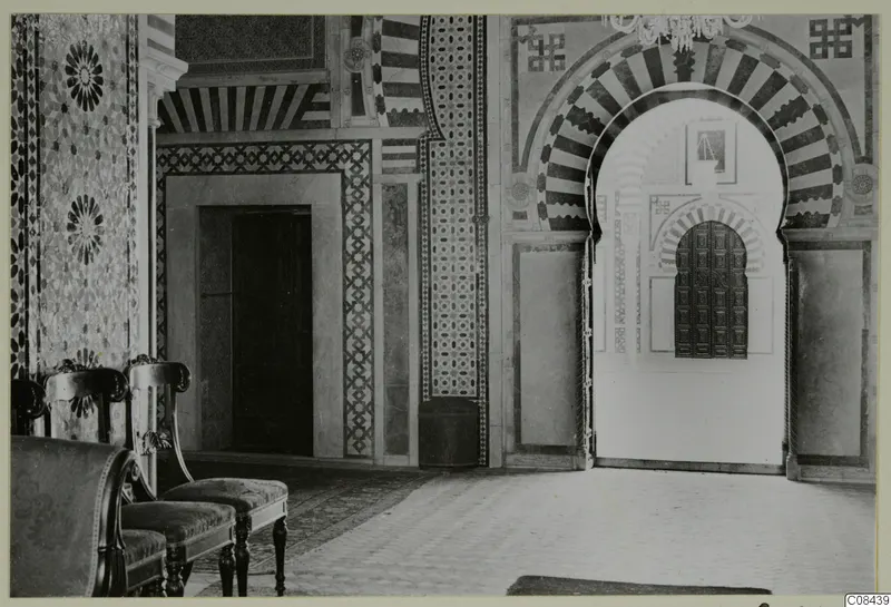 Interior of a building with a striped arch framing a studded wooden door, patterned wall panels and upholstered chairs in the foreground.