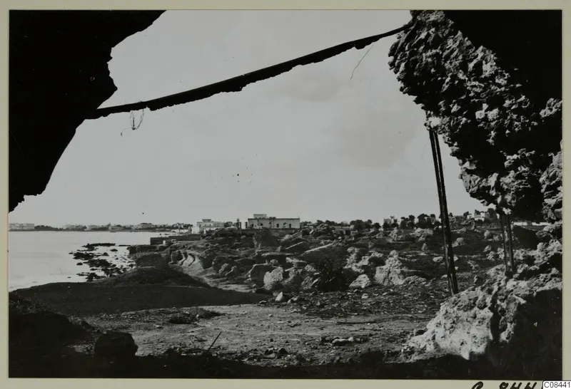 Rocky coastline and stones seen from under a dark rock overhang, with water and buildings on the horizon, North Africa.