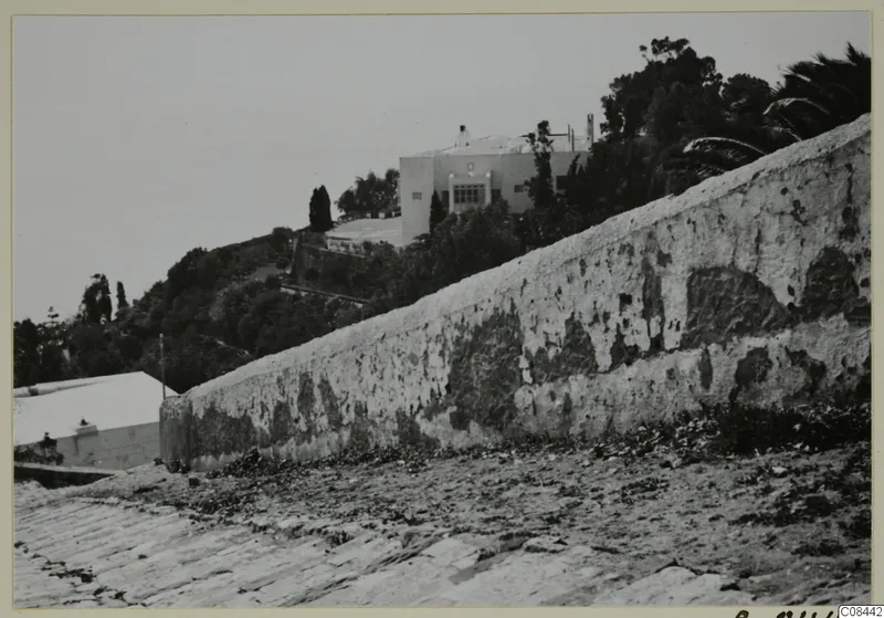 Weathered plaster wall running up a slope, with a white building and trees behind, North Africa