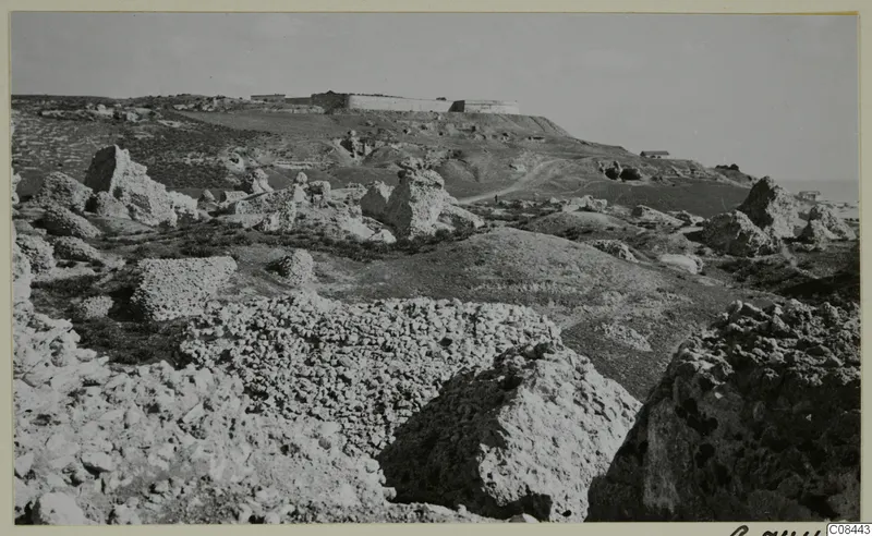 Rocky limestone ground with fallen stone blocks, a long walled structure on a hilltop and a small building, with the sea at right.