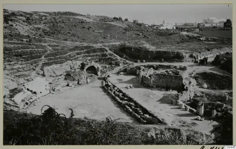 Ruined stone walls and a vaulted arch on a hillside site, with a white domed building in the distance