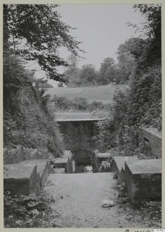 Stone-walled steps leading up a gravel path toward a brick-faced structure, with trees and bushes on both sides