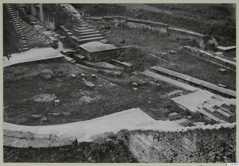 Excavated brick ruins with stepped brick structures, low walls and paved surfaces on a grassy site, seen from above