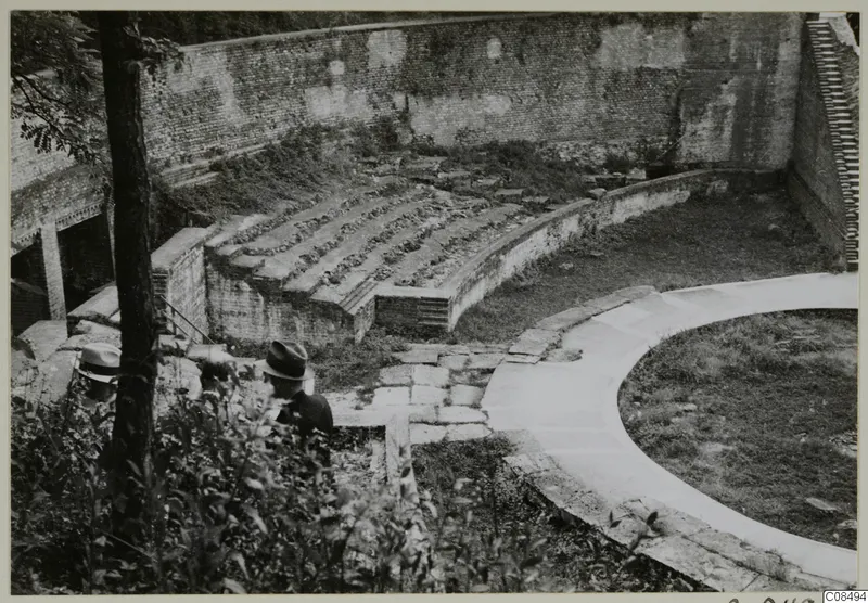 Curved brick-walled structure with tiered stone seating and a paved walkway, with two men in hats at the left edge