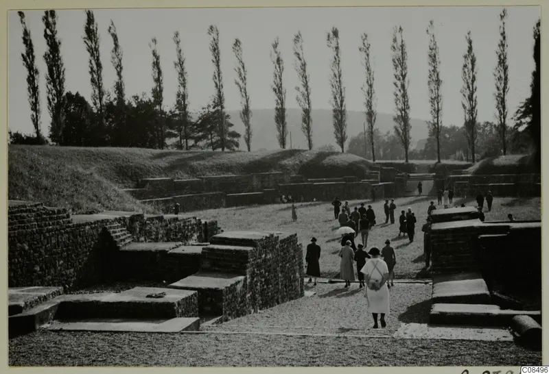 Visitors walking among stone ruins and low walls, with a grassy bank and tall poplars behind.