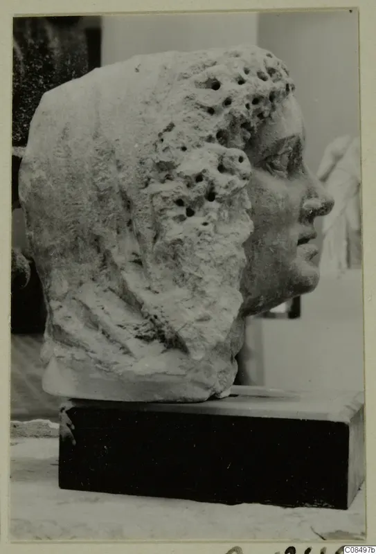 Carved stone head in profile facing right, with curly hair pitted by drill holes, on a dark rectangular base
