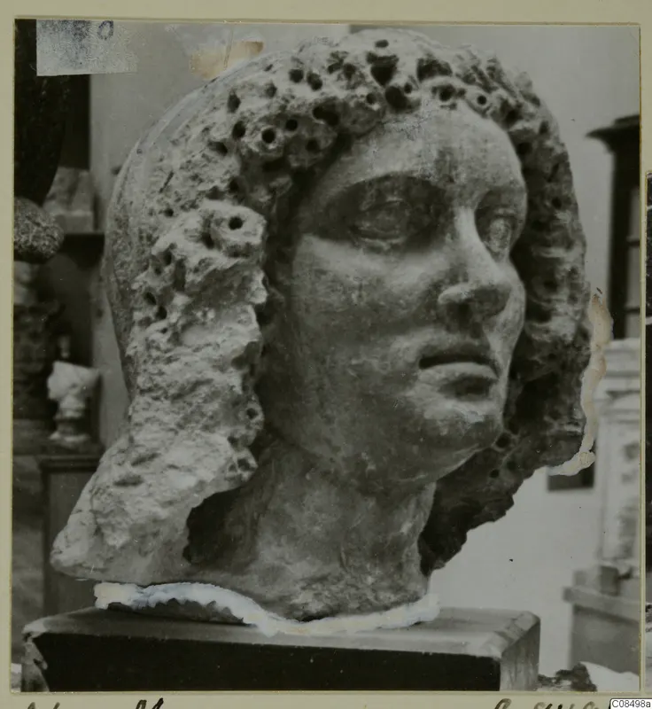 Sculpted stone head with curled hair pierced by many small holes, on a dark plinth